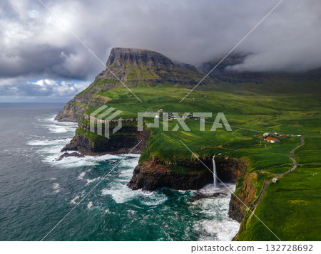 Dramatic Mulafossur waterfall plunging into the ocean with Gasadalur village and cliffs in the background. 132728692