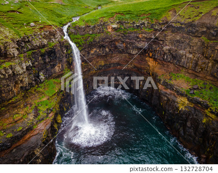 Dramatic Mulafossur waterfall plunging into the ocean with Gasadalur village and cliffs in the background. 132728704