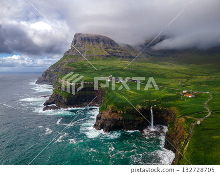 Dramatic Mulafossur waterfall plunging into the ocean with Gasadalur village and cliffs in the background. 132728705