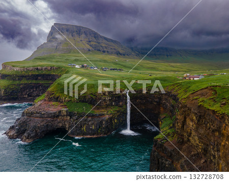 Dramatic Mulafossur waterfall plunging into the ocean with Gasadalur village and cliffs in the background. Dramatic Mulafossur waterfall plunging into the ocean with Gasadalur village and cliffs in the background. 132728708