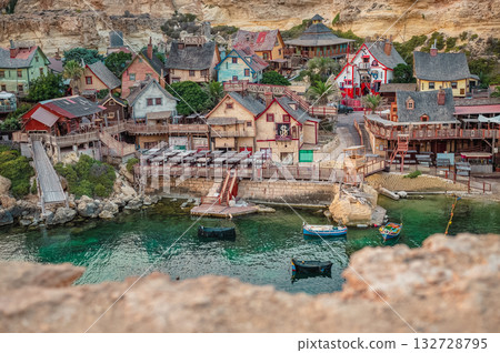 A picturesque view of the famous Popeye Village and its beautiful bay in Malta, with colorful wooden houses and turquoise waters shining under the bright sun. A picturesque view of the famous Popeye Village and its beautiful bay in Malta, with colorful wooden houses and turquoise waters shining under the bright sun. 132728795