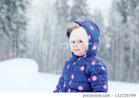 Outdoor winter portrait of little cute toddler girl in blue coat and white fashion hat. Healthy happy baby child playing in the park on cold day with snow and snowfall. Stylish clothes for kids 132728799