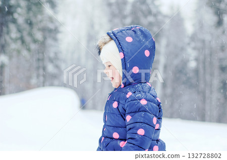 Outdoor winter portrait of little cute toddler girl in blue coat and white fashion hat. Healthy happy baby child playing in the park on cold day with snow and snowfall. Stylish clothes for kids Outdoor winter portrait of little cute toddler girl in blue coat and white fashion hat. Healthy happy baby child playing in the park on cold day with snow and snowfall. Stylish clothes for kids 132728802