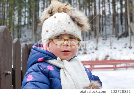 Outdoor winter portrait of little cute toddler girl in blue coat and white fashion hat. Healthy happy baby child playing in the park on cold day with snow and snowfall. Stylish clothes for kids Outdoor winter portrait of little cute toddler girl in blue coat and white fashion hat. Healthy happy baby child playing in the park on cold day with snow and snowfall. Stylish clothes for kids 132728821