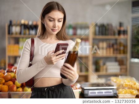 European young woman customer at shop scans QR code on beer glass bottle using phone camera 132730356