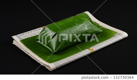 Banana Leaf Wrapped Food on Newspaper Against Black Background Banana Leaf Wrapped Food on Newspaper Against Black Background 132730364