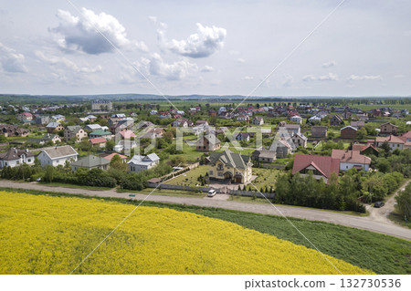 Aerial view of ground road in green fields with blooming rapeseed plants, suburb houses on horizon and blue sky copy space background. Drone photography. Aerial view of ground road in green fields with blooming rapeseed plants, suburb houses on horizon and blue sky copy space background. Drone photography. 132730536