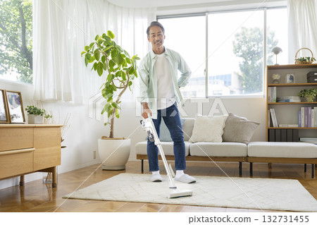 一位六十多歲的男子正在家裡的客廳裡用吸塵器打掃衛生,他看著鏡頭。 一位六十多歲的男子正在家裡的客廳裡用吸塵器打掃衛生,他看著鏡頭。 132731455