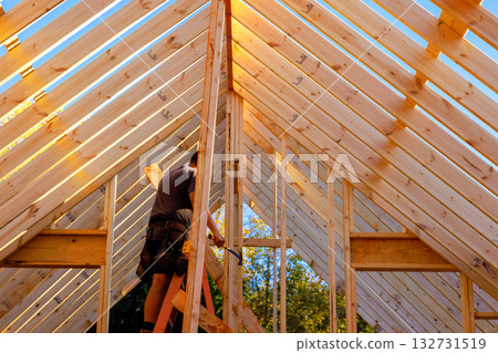 Worker constructs wooden beams frame under on roof structure of new building. Worker constructs wooden beams frame under on roof structure of new building. 132731519