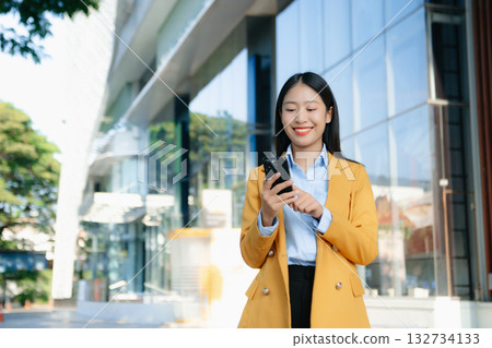 Confident Asian businesswoman smiling while using a smartphone outdoors, symbolizing technology, success, and modern professional lifestyle. 132734133