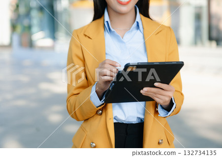 Confident Asian businesswoman smiling while using a smartphone outdoors, symbolizing technology, success, and modern professional lifestyle. 132734135