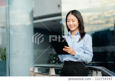 Asian woman with a smile standing holding notepad and tablet at out side office. big city on business center 132734147