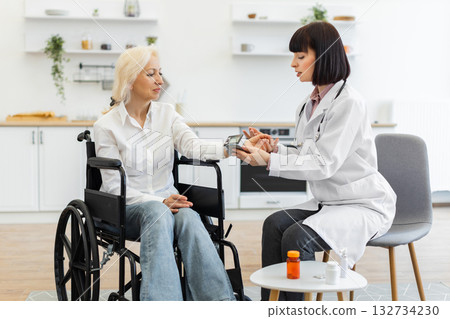A female doctor measures the blood pressure of an elderly woman in a wheelchair during a home visit, with medicine on a table. 132734230