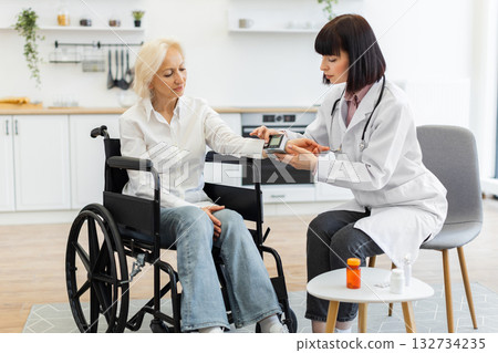 A female doctor in a white coat checks the blood pressure of an elderly woman seated in a wheelchair, indoors. 132734235