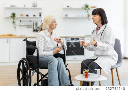 A female doctor in a white coat explains medication to an elderly woman in a wheelchair, discussing treatment options in a home setting. 132734240