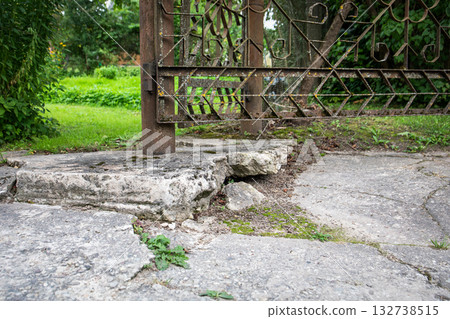 A weathered metal gate attached to a rusty pole standing on broken and crumbling concrete base in a 132738515