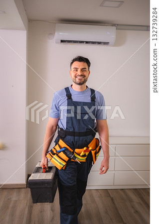 Technician smiling carrying toolbox and wearing tool belt with air conditioner in background 132739284