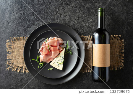Bottle of red wine with blue cheese, prosciutto, and rosemary on a black table. Bottle of red wine with blue cheese, prosciutto, and rosemary on a black table. 132739675