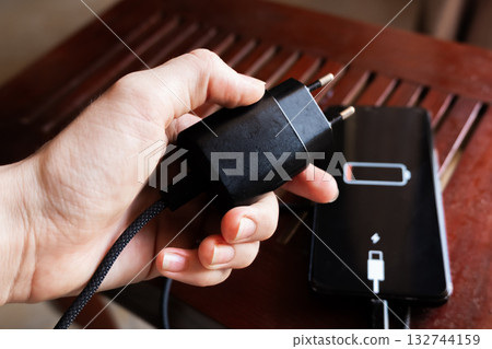 A black smartphone with a low battery icon is connected to a charger on a wooden surface. The charger is plugged into a power outlet. A black smartphone with a low battery icon is connected to a charger on a wooden surface. The charger is plugged into a power outlet. 132744159