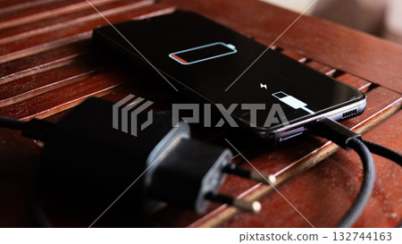 A black smartphone with a low battery icon is connected to a charger on a wooden surface. The charger is plugged into a power outlet. A black smartphone with a low battery icon is connected to a charger on a wooden surface. The charger is plugged into a power outlet. 132744163