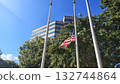 A half-masted American flag flutters in a light breeze against a backdrop of a brown office or government building and green trees, the US national flag at half-mast on a city street on a sunny day 132744864