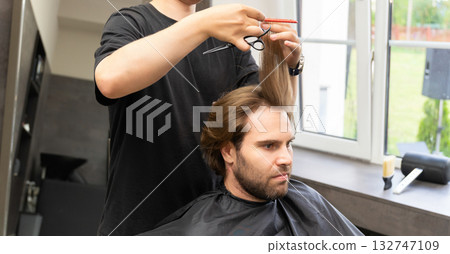 A barber uses scissors and a comb to give a haircut to a male client with long brown hair. 132747109