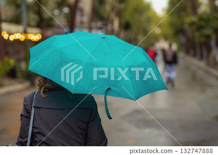 Woman back view with green umbrella walking on rainy city street surrounded by soft bokeh lights and blurred background Woman back view with green umbrella walking on rainy city street surrounded by soft bokeh lights and blurred background 132747888