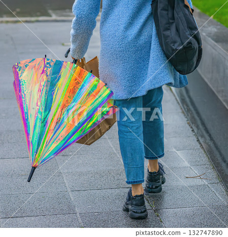 Person walking on wet pavement holding folded iridescent umbrella with bright rainbow reflections. Modern street fashion, rainy weather and vibrant urban lifestyle 132747890