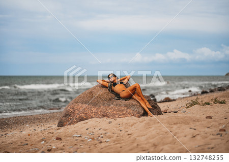 A woman enjoys a serene moment on the beach, lounging against a rock as waves gently lap at her feet A woman enjoys a serene moment on the beach, lounging against a rock as waves gently lap at her feet 132748255
