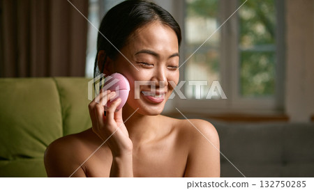 Smiling woman massaging face with pink cleansing device at home Smiling woman massaging face with pink cleansing device at home 132750285