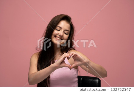 Happy brunette woman with long hair smiling and looking down making heart sign on pink background Happy brunette woman with long hair smiling and looking down making heart sign on pink background 132752547