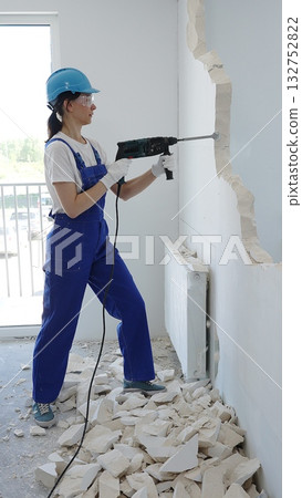 Female construction worker demolishing interior wall with rotary hammer, wearing safety glasses and hardhat, creating substantial concrete debris pile during demolition process 132752822
