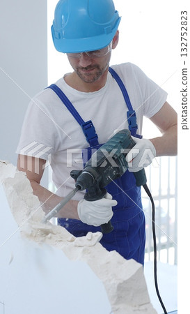 Male construction worker demolishing concrete wall with rotary hammer, wearing protective safety overalls is generating dust and debris during renovation project. Vertical portrait view 132752823
