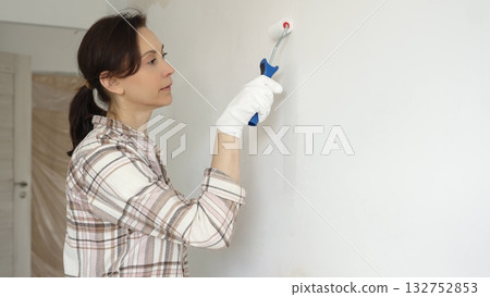 Woman wearing protective gloves painting white wall with small paint roller, updating interior and giving room clean, modern look during home renovation project Woman wearing protective gloves painting white wall with small paint roller, updating interior and giving room clean, modern look during home renovation project 132752853