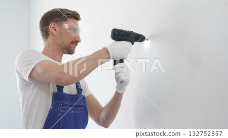Middle aged man wearing blue overall and protective glasses, is drilling hole in white wall, wearing protective gear during home improvement project with cordless power tool 132752857