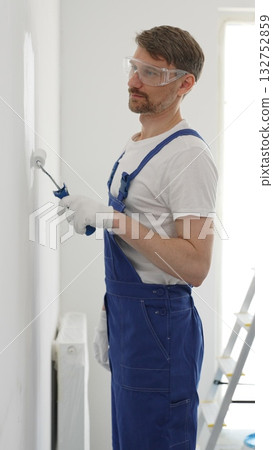 Professional painter wearing protective white gloves and blue work overalls, applying paint with roller on pristine interior wall during home renovation project Professional painter wearing protective white gloves and blue work overalls, applying paint with roller on pristine interior wall during home renovation project 132752859