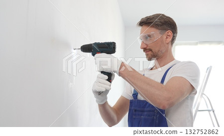 Man professional construction worker in a blue jumpsuit, safety goggles, and work gloves is screwing a screw into the wall using a cordless screwdriver, home renovation 132752862