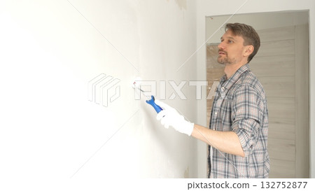 Man professional painter wearing checkered shirt, is rolling white paint on apartment wall with small blue roller, transforming interior space with steady strokes during comprehensive home renovation Man professional painter wearing checkered shirt, is rolling white paint on apartment wall with small blue roller, transforming interior space with steady strokes during comprehensive home renovation 132752877