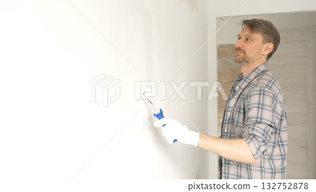 Man professional painter wearing checkered shirt, is rolling white paint on apartment wall with small blue roller, transforming interior space with steady strokes during comprehensive home renovation Man professional painter wearing checkered shirt, is rolling white paint on apartment wall with small blue roller, transforming interior space with steady strokes during comprehensive home renovation 132752878