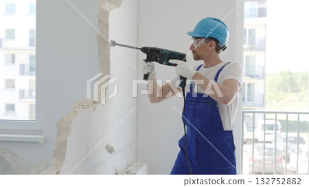 Male construction worker demolishing concrete wall with rotary hammer, wearing work overalls and blue hardhat is generating dust and debris during home renovation work, portrait horizontal view 132752882