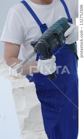 Construction worker demolishing white wall with rotary hammer drill, wearing blue overalls and protective white gloves, generating dust during renovation project, closeup view 132752886