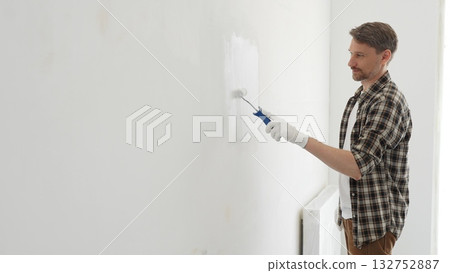 Middle aged male painter wearing plaid shirt and protective gloves, is painting a white wall with a paint roller, working on a home renovation project, creating a fresh and clean look Middle aged male painter wearing plaid shirt and protective gloves, is painting a white wall with a paint roller, working on a home renovation project, creating a fresh and clean look 132752887