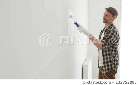 Middle aged male painter wearing plaid shirt and protective gloves, is painting a white wall with a paint roller, working on a home renovation project, creating a fresh and clean look Middle aged male painter wearing plaid shirt and protective gloves, is painting a white wall with a paint roller, working on a home renovation project, creating a fresh and clean look 132752893