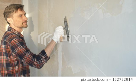 Male construction worker wearing protective gloves smoothing plaster onto wall surface during home renovation using professional trowel tool 132752914