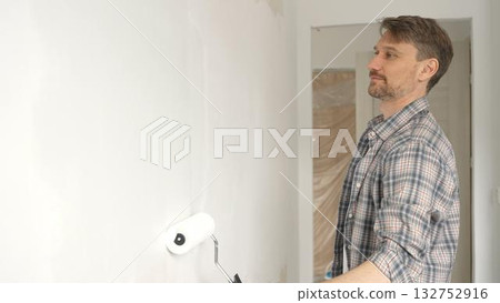Middle aged man wearing checkered shirt, is rolling white paint on apartment wall with paint roller, transforming interior space with steady strokes during comprehensive home renovation Middle aged man wearing checkered shirt, is rolling white paint on apartment wall with paint roller, transforming interior space with steady strokes during comprehensive home renovation 132752916