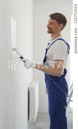 Male professional painter wearing protective white gloves and blue work overalls, applying paint with roller on pristine interior wall during home renovation project, vertical photo Male professional painter wearing protective white gloves and blue work overalls, applying paint with roller on pristine interior wall during home renovation project, vertical photo 132752919