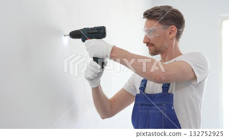 Professional handyman wearing blue overall, safety glasses and gloves, is using cordless drill to make hole in white wall during renovation or installation work 132752925