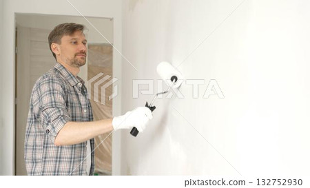 Man professional painter wearing checkered shirt, is rolling white paint on apartment wall with paint roller, transforming interior space with steady strokes during comprehensive home renovation Man professional painter wearing checkered shirt, is rolling white paint on apartment wall with paint roller, transforming interior space with steady strokes during comprehensive home renovation 132752930