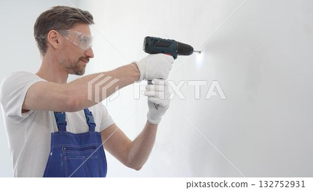 Middle aged man wearing blue overall, is drilling hole in white wall, wearing protective gear during home improvement project with cordless power tool 132752931