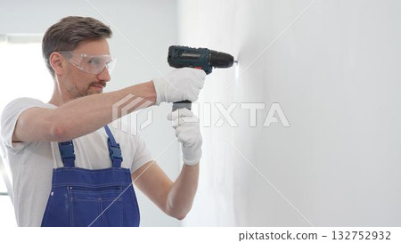 Professional handyman wearing blue overall, safety glasses and gloves, is using cordless drill to make hole in white wall during renovation or installation work 132752932